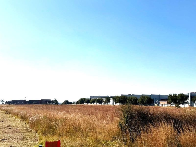 A wide-angle view of a vacant commercial land plot featuring dry grass and surrounding industrial or commercial developments in the distance.