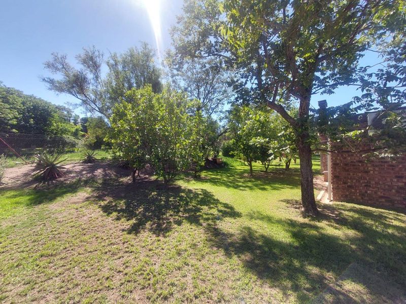 A spacious, established garden area featuring mature trees, lush lawn, and a partial view of the brick exterior wall of the farm dwelling.