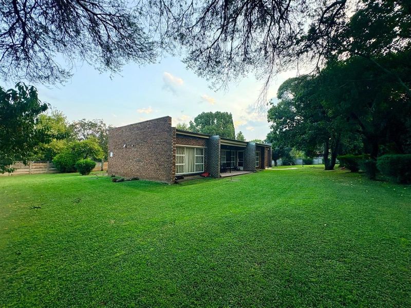 A spacious rear garden featuring a well-maintained lawn, mature trees, and a face brick house with a covered patio and sliding glass doors.