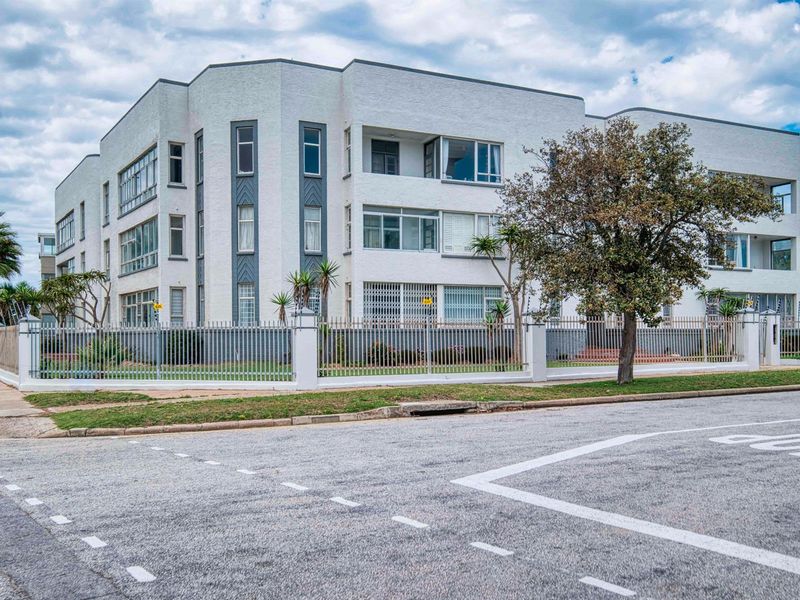 A street-level view of a multi-story apartment building featuring a white facade, metal perimeter fencing, and established landscaping.
