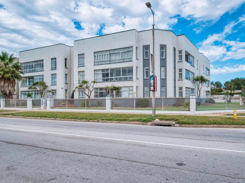 Street-facing view of a multi-story apartment building featuring a white concrete facade, metal perimeter fencing, and enclosed balcony sections.