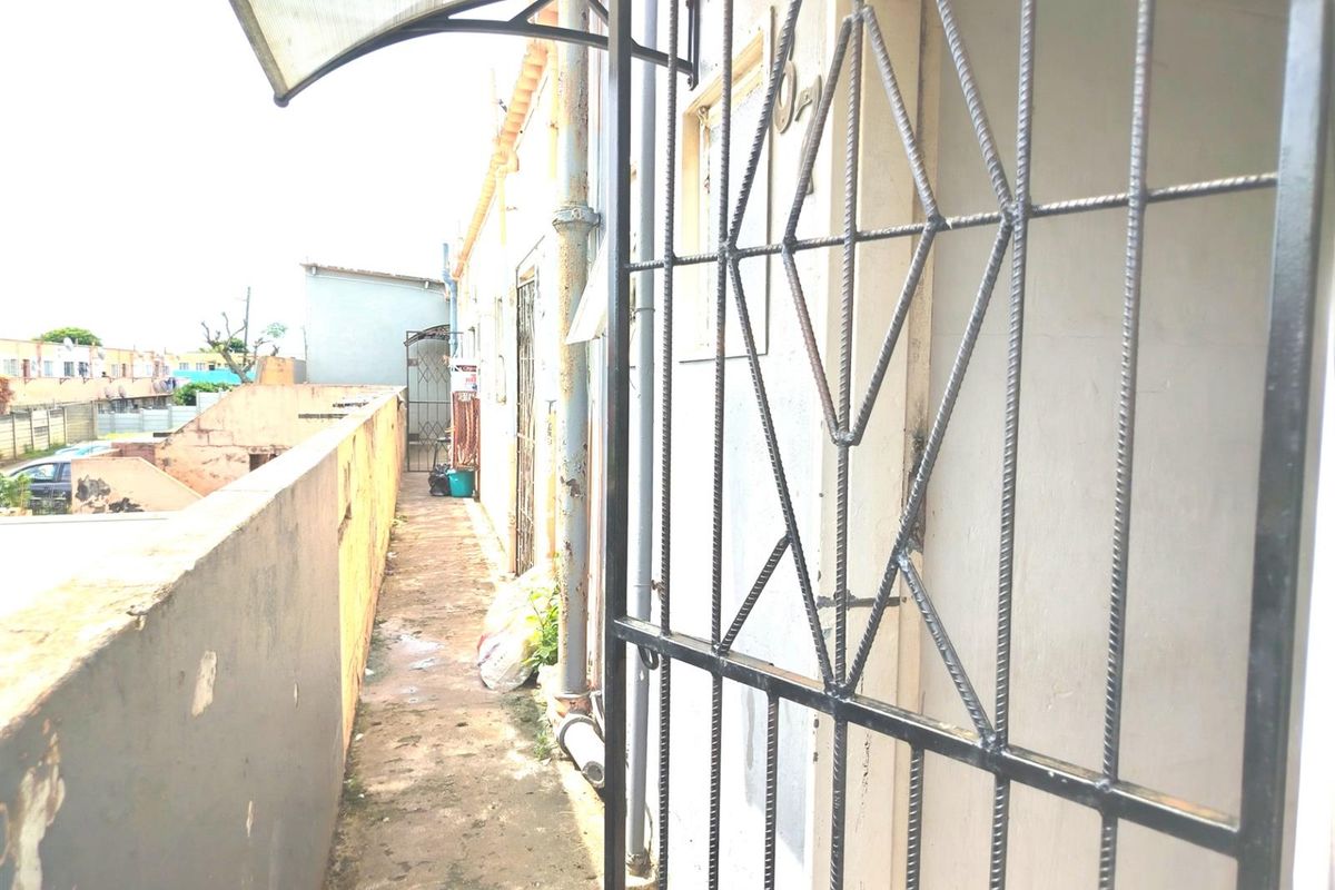 A narrow exterior rear walkway featuring a security gate, protective awning, and weathered boundary wall with visible signs of wear and surface deterioration.