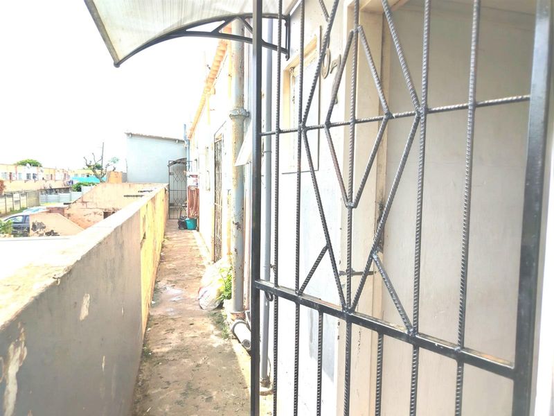A narrow exterior rear walkway featuring a security gate, protective awning, and weathered boundary wall with visible signs of wear and surface deterioration.