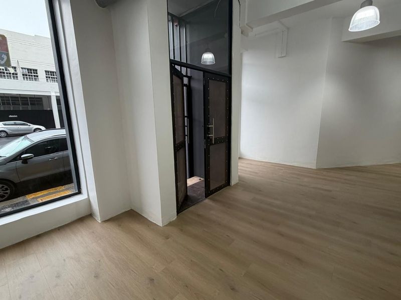 A bright, open-plan retail space featuring modern wood-look flooring, large display windows, and a secure industrial-style entrance door.