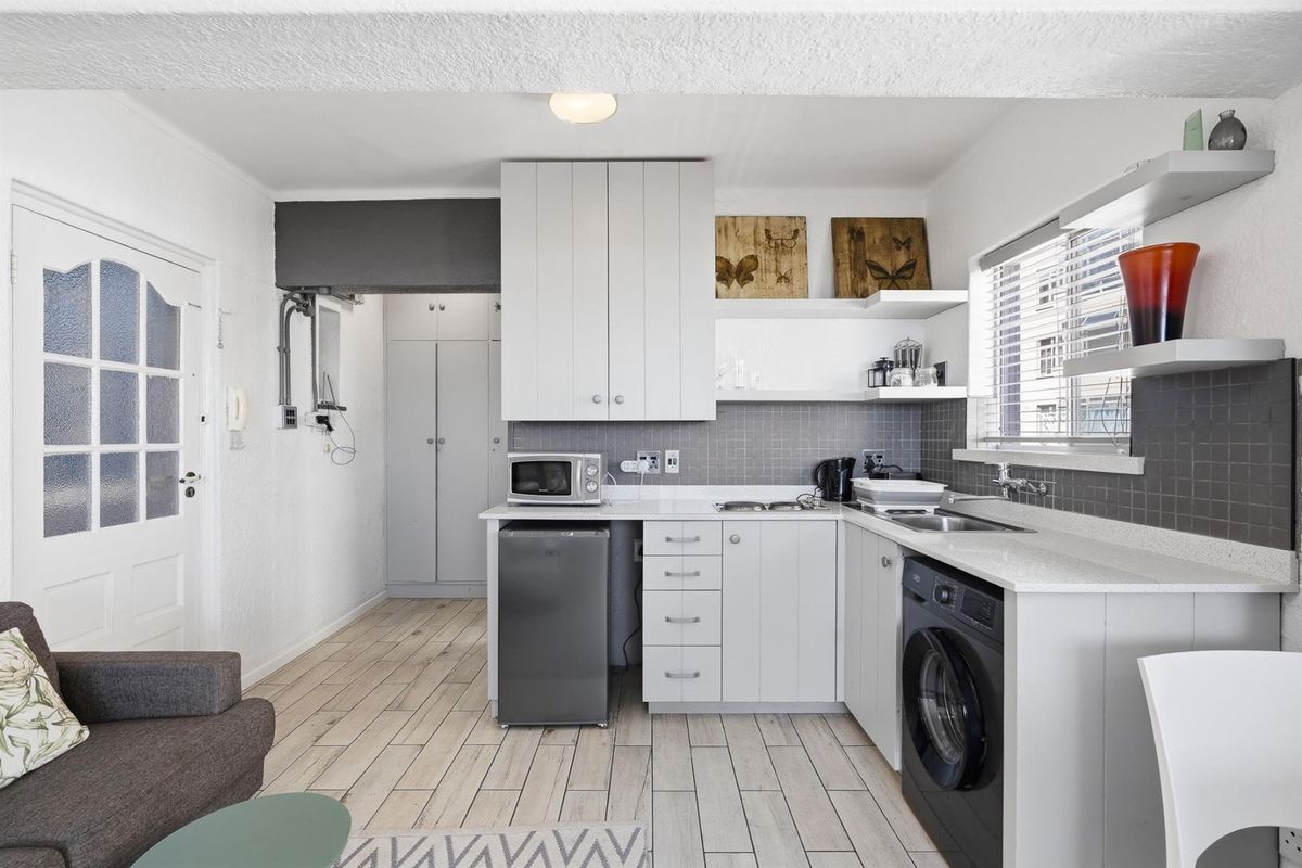 Compact kitchen featuring white cabinetry, grey tiled backsplash, integrated sink, and space for a washing machine, with light wood-look flooring throughout.
