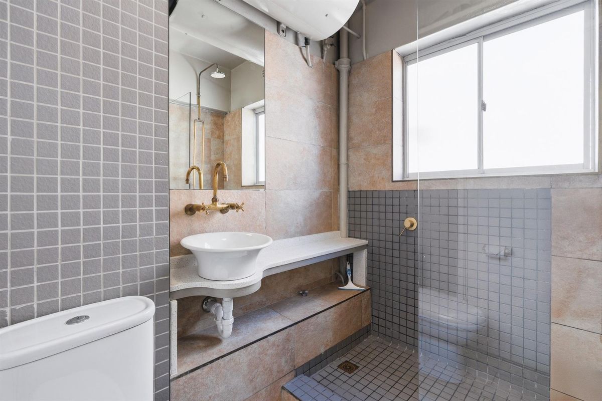 Compact bathroom featuring a vessel sink, wall-mounted brass faucet, glass shower screen, and a mix of grey and beige mosaic wall tiling.