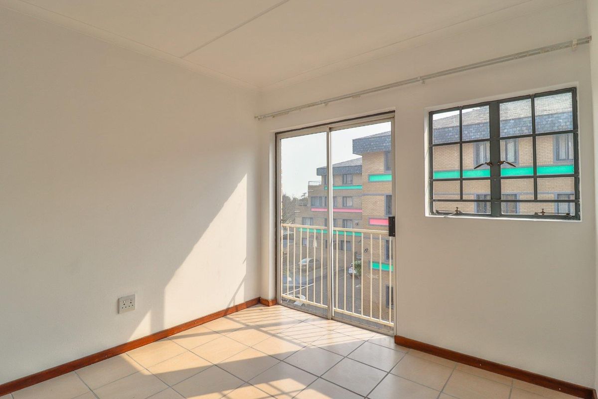 A bright, empty bedroom featuring tiled flooring, a sliding glass door leading to a balcony, and a window with views of the surrounding apartment complex.