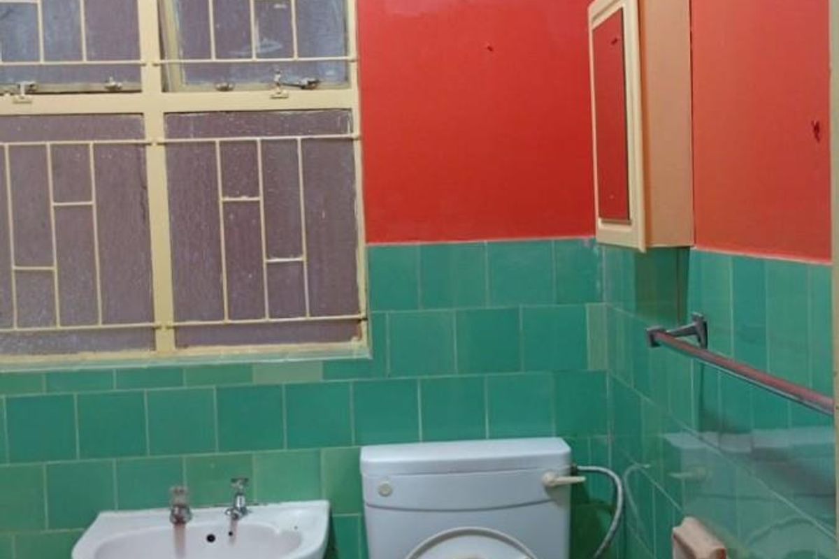A dated bathroom featuring green wall tiles, a white pedestal basin, a standard toilet, and a metal-framed window with security bars.