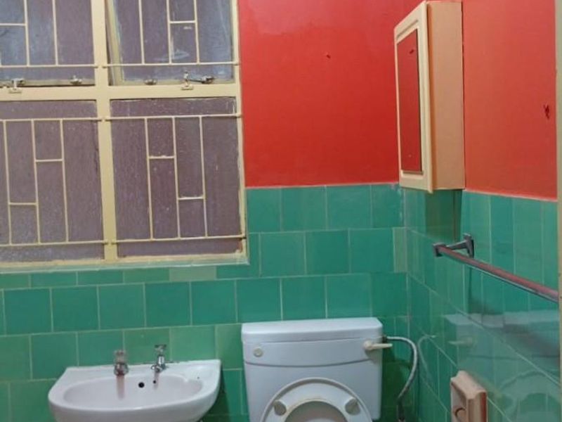 A dated bathroom featuring green wall tiles, a white pedestal basin, a standard toilet, and a metal-framed window with security bars.