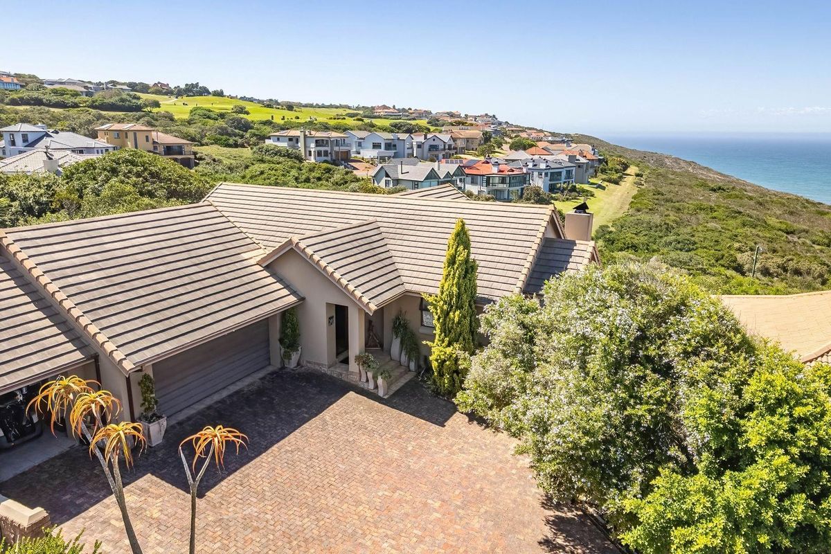 Aerial view of a coastal residence featuring a tiled roof, paved driveway, and double garage, set within a scenic neighbourhood overlooking the ocean.