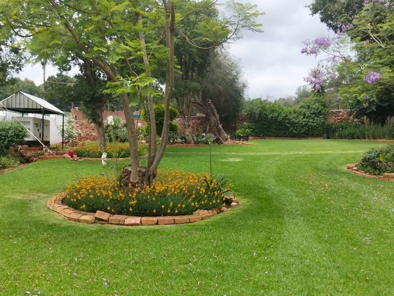 A well-maintained garden area featuring established trees, landscaped flower beds with brick edging, and a spacious lawn on a small holding property.