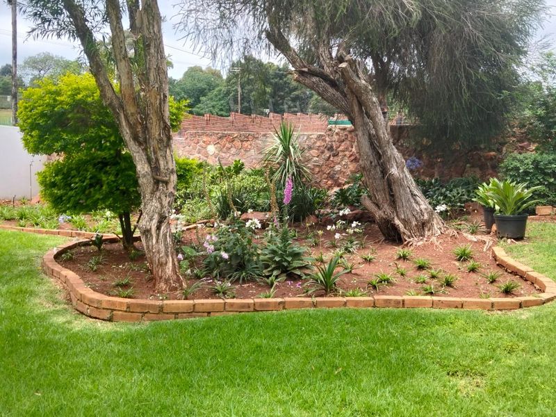 A well-maintained garden area featuring established trees, landscaped flower beds with brick edging, and a stone boundary wall.