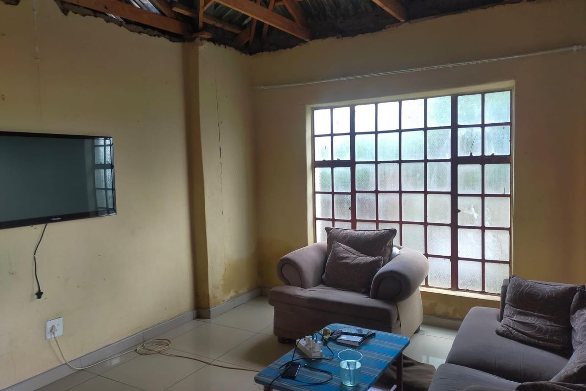 Living room featuring tiled flooring, large grid-pattern window, and exposed timber roof trusses, requiring significant renovation and ceiling installation.