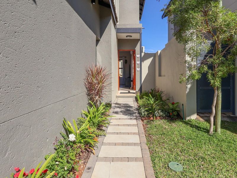 A paved walkway leads to the front entrance of the townhouse, flanked by landscaped garden beds and boundary walls.