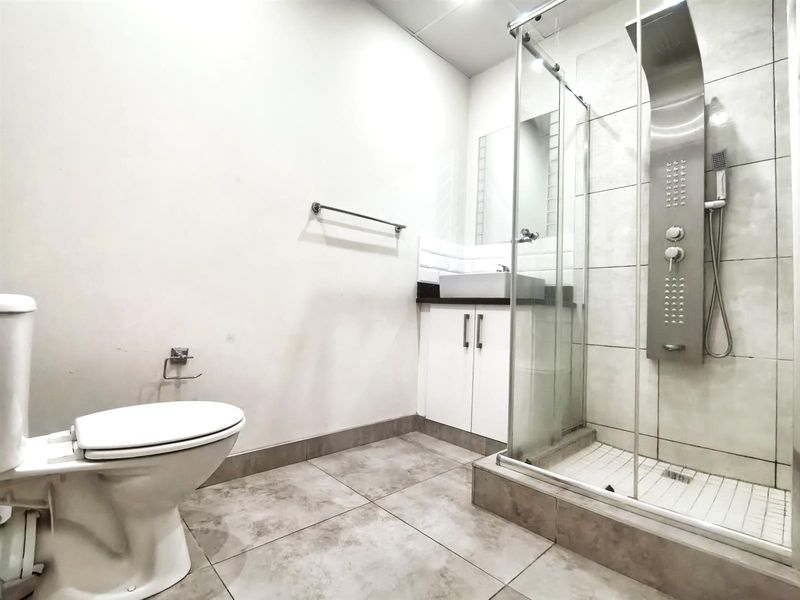 A clean, modern bathroom featuring a glass-enclosed shower with a stainless steel shower panel, a vessel sink vanity, and neutral floor tiling.