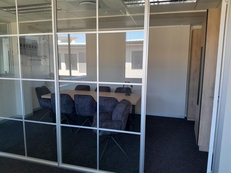 A modern office meeting room featuring a glass partition wall, suspended ceiling, and built-in cabinetry with dark carpet flooring.