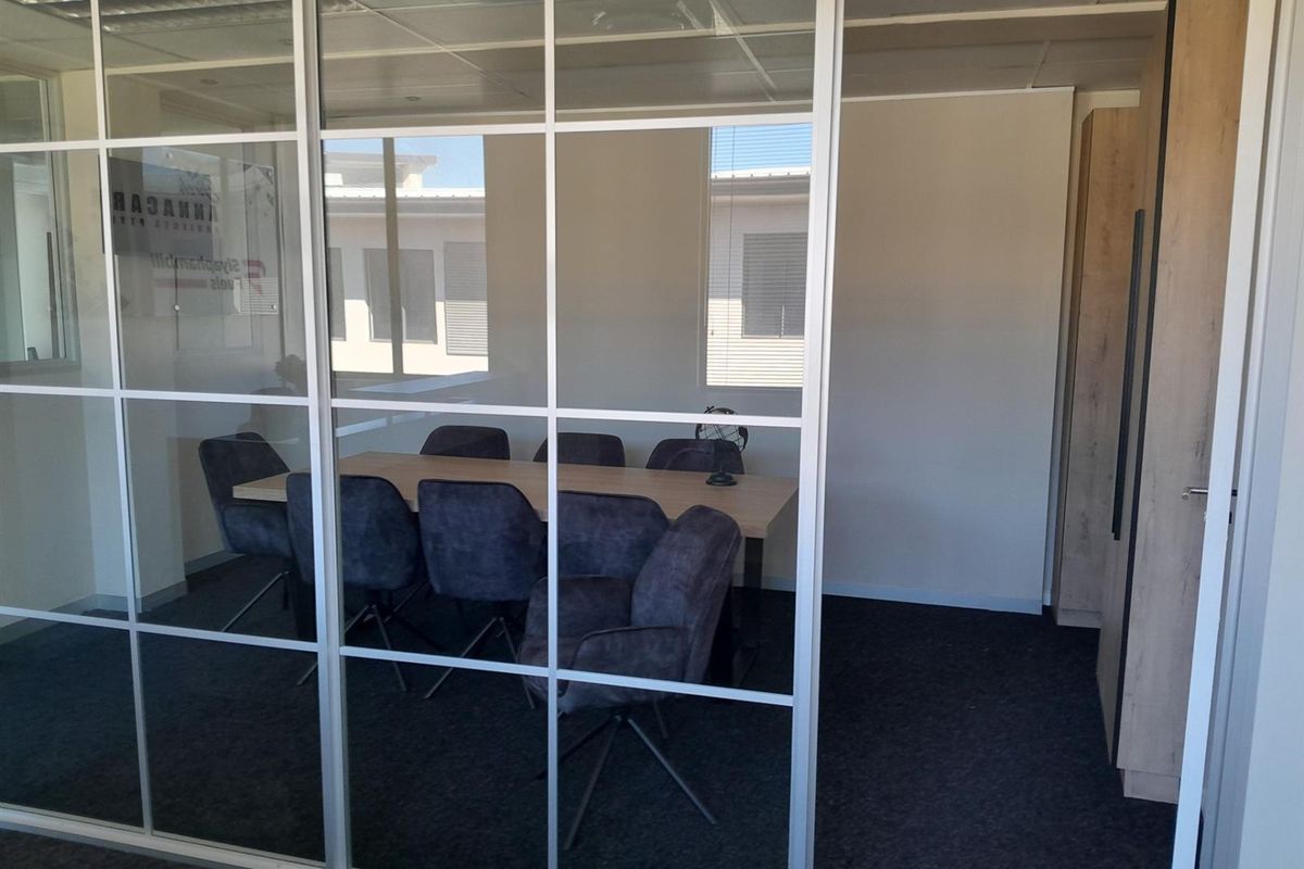 A modern office meeting room featuring a glass partition wall, suspended ceiling, and built-in cabinetry with dark carpet flooring.