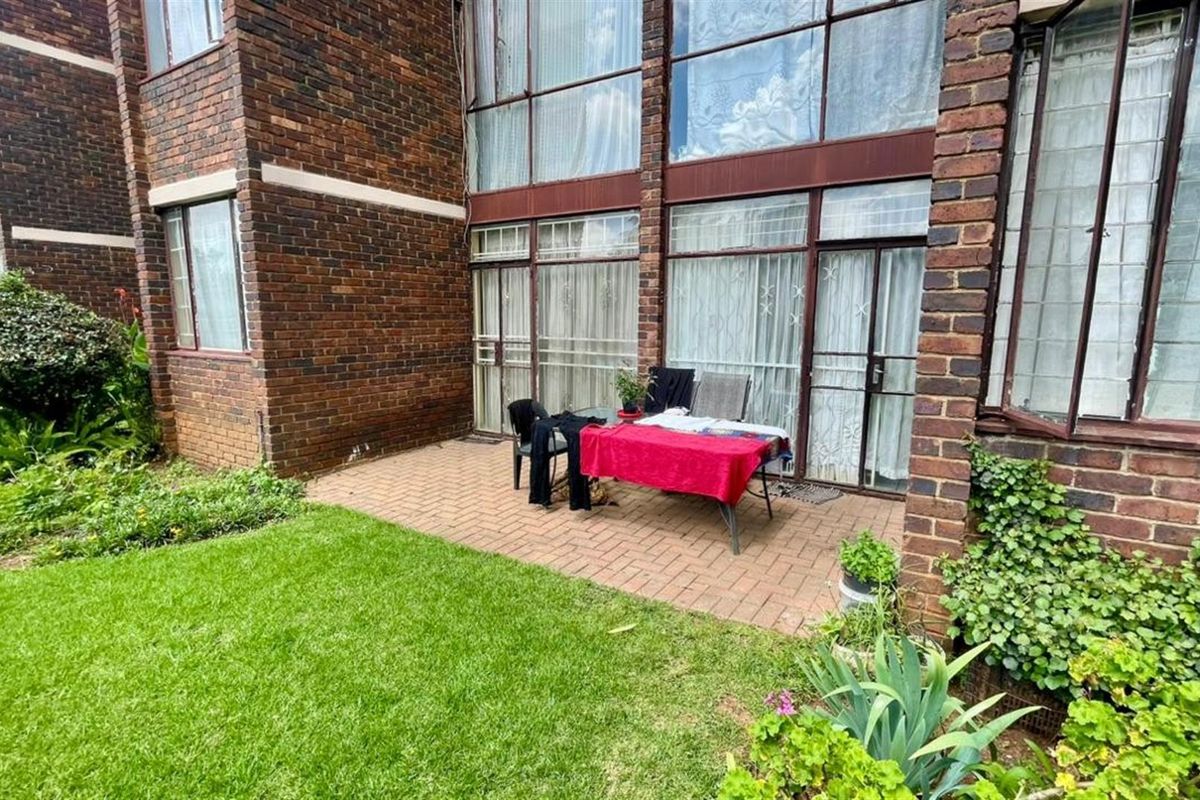 A rear view of the apartment featuring a paved patio area, established garden, and large floor-to-ceiling windows with brick exterior walls.