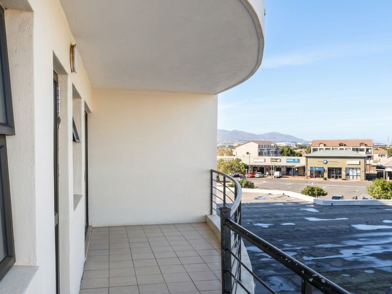 A covered balcony with tiled flooring and a metal railing, offering an elevated view of the surrounding commercial area and distant mountains.