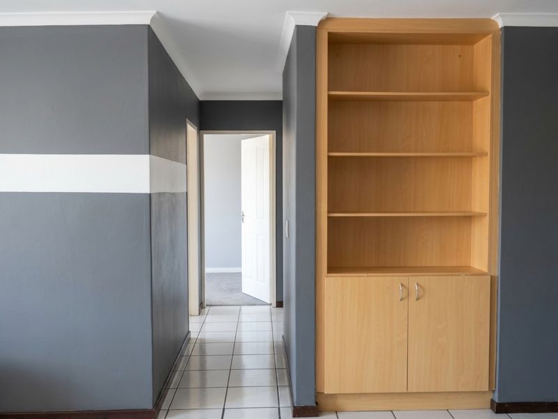 A hallway featuring tiled flooring, a built-in wooden shelving unit, and access to an adjacent room with carpeted flooring.