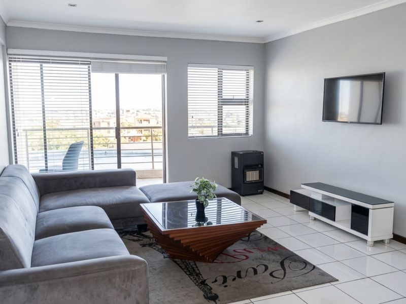 A bright living room featuring tiled flooring, large windows with blinds, and access to a balcony, creating a comfortable and functional living space.