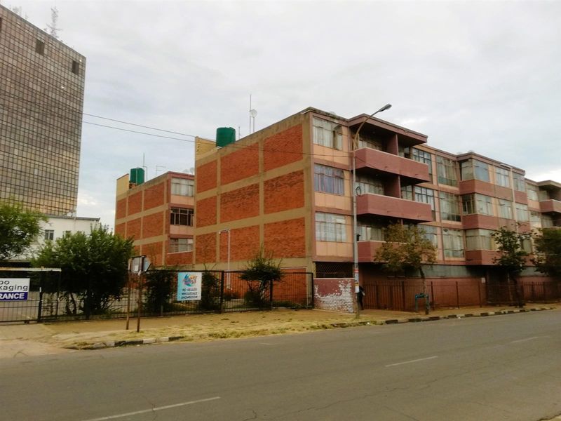Street view of a multi-story apartment building featuring a brick facade, integrated balconies, and perimeter fencing.
