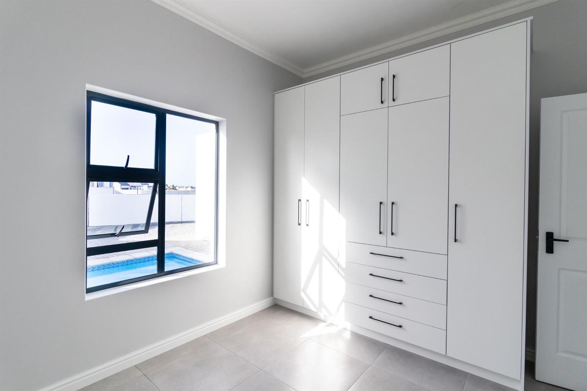 A bright, modern bedroom featuring sleek white built-in cupboards, neutral tiled flooring, and a window view overlooking the swimming pool area.