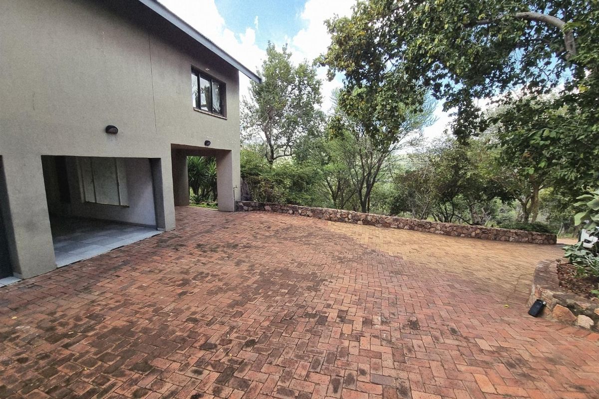 Rear exterior view featuring a brick paved driveway, stone retaining wall, and a covered parking area surrounded by mature trees.