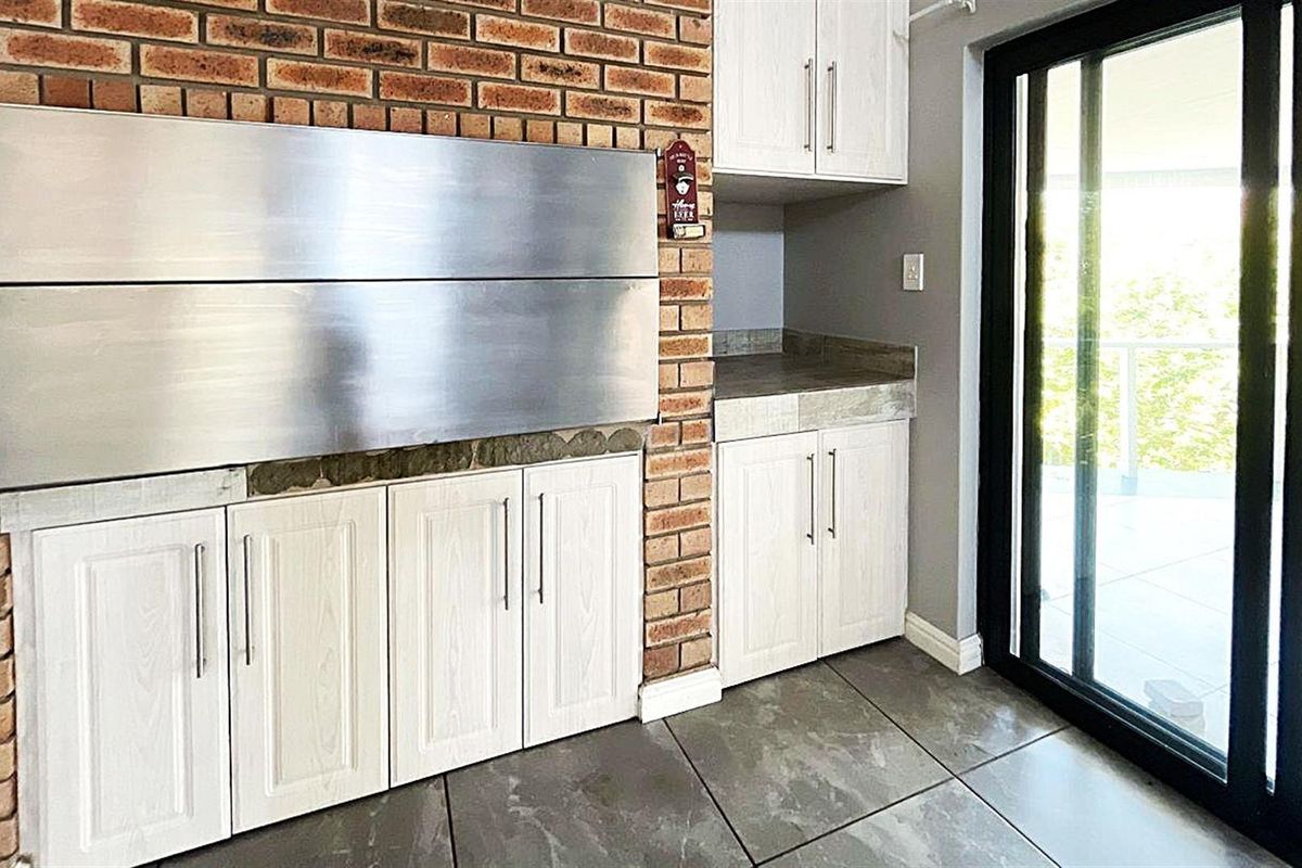Indoor entertainment area featuring a built-in stainless steel braai, white cabinetry, stone-look countertops, and large format floor tiles with sliding door access to the patio.