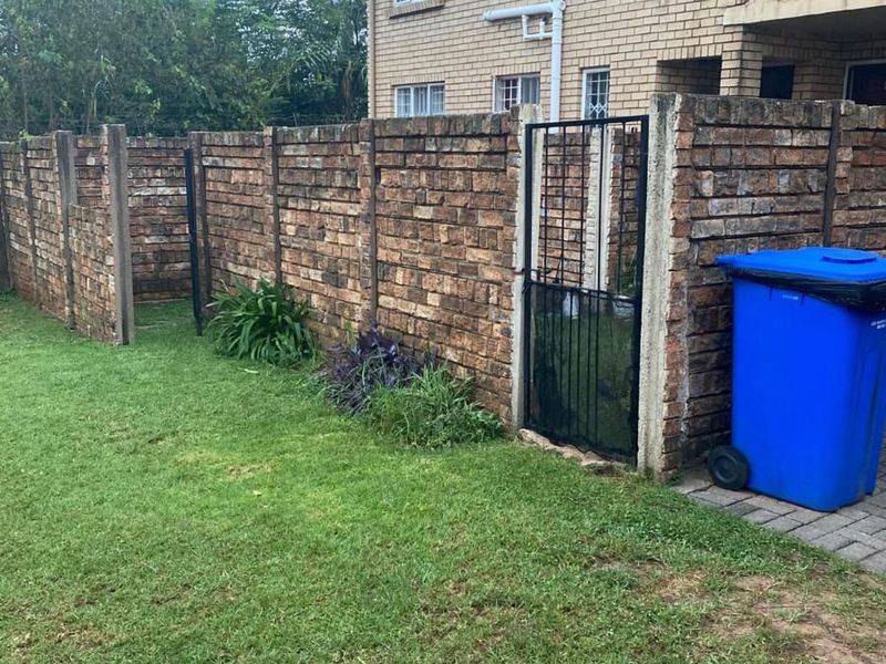 Rear exterior view of the apartment building featuring a face brick boundary wall, metal pedestrian gate, paved walkway, and a private lawn area.