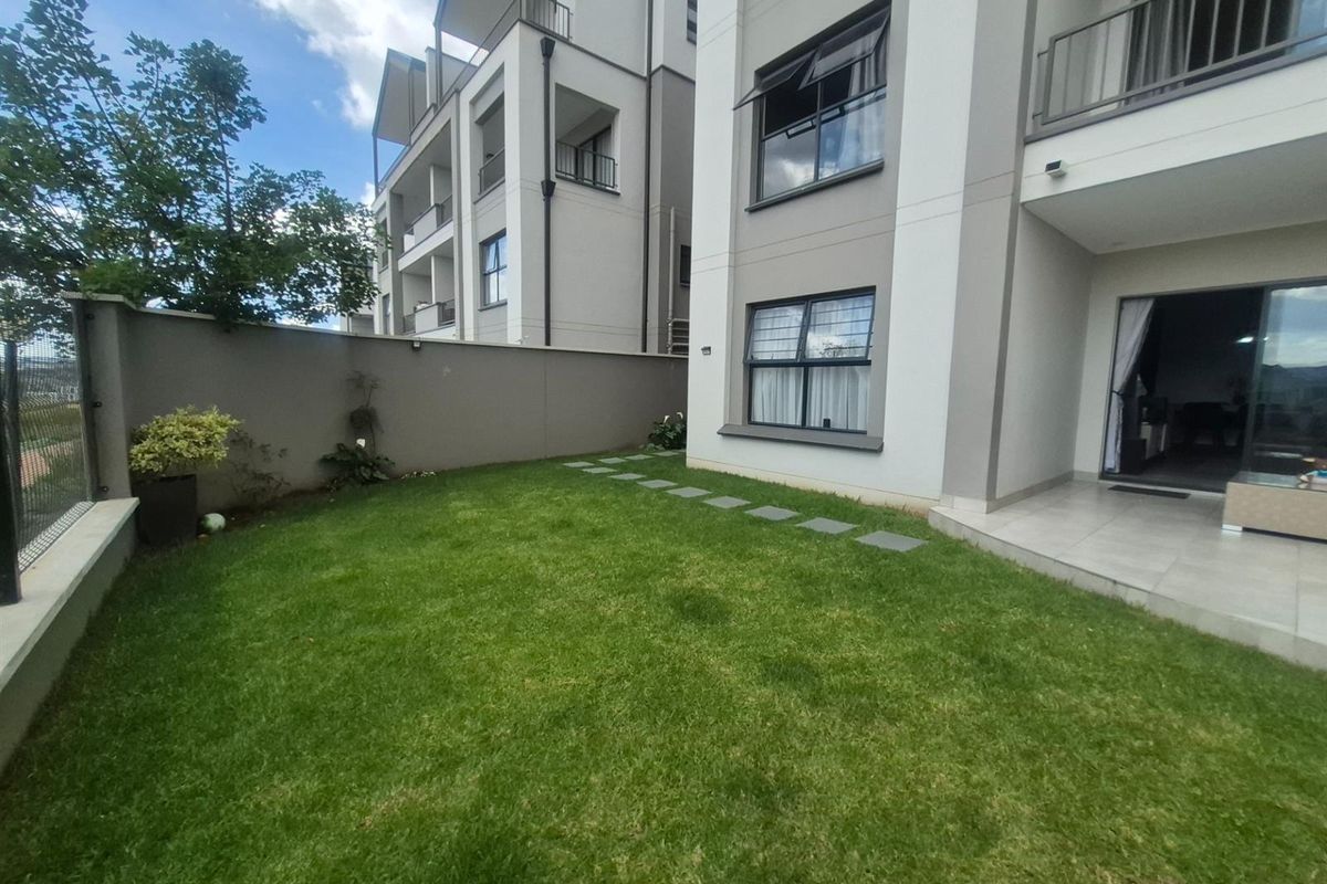 A private rear garden area featuring a manicured lawn, paved stepping stones, and a tiled patio area adjacent to the apartment building.