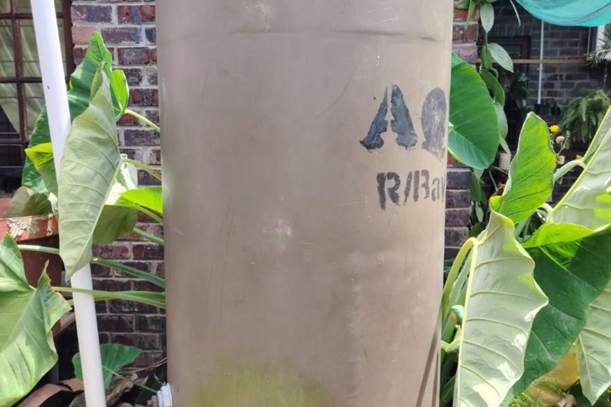 An exterior view of a water storage tank positioned against a brick wall, featuring connected plumbing infrastructure.