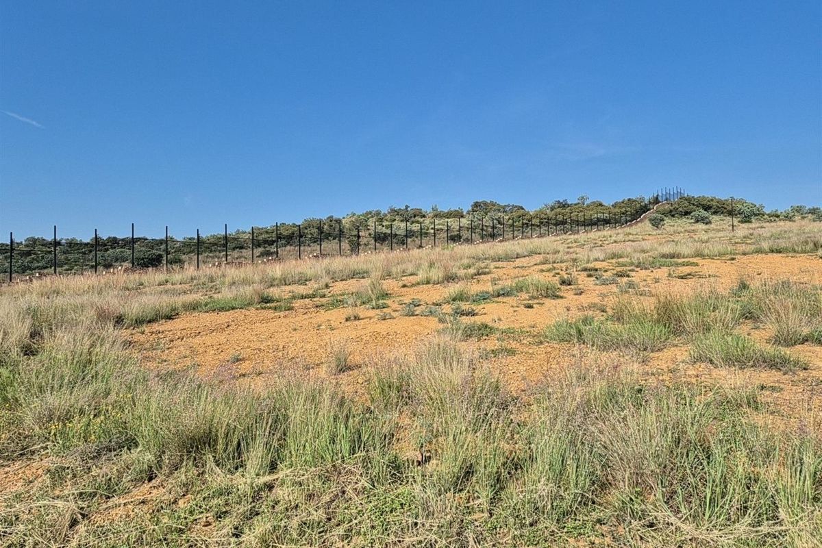 A view of a vacant residential plot featuring natural terrain and perimeter fencing against a clear blue sky.