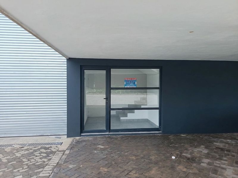 Front entrance of an industrial property featuring a modern glass door, metal roller shutter, and paved driveway under a covered canopy.