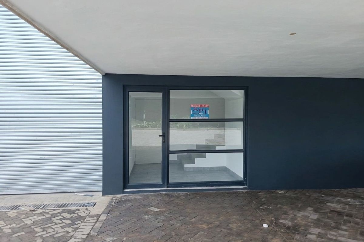 Front entrance of an industrial property featuring a modern glass door, metal roller shutter, and paved driveway under a covered canopy.