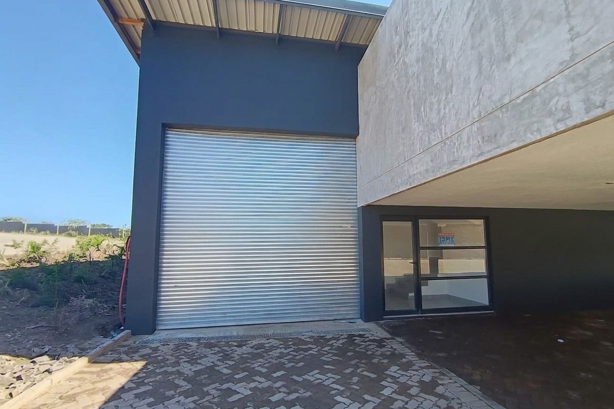 Modern industrial facade featuring a large metal roller shutter door, glass entrance, and paved driveway area.