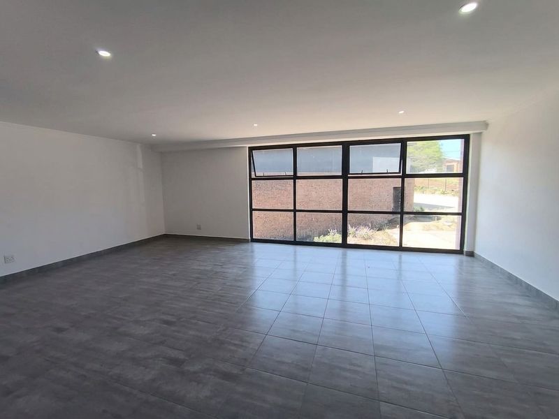 A spacious, empty open-plan area featuring modern grey tiled flooring, recessed downlights, and large black-framed windows providing ample natural light.