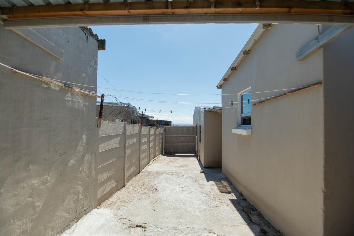 A narrow rear exterior passage featuring a concrete driveway, boundary wall with security wire, and a timber gate.