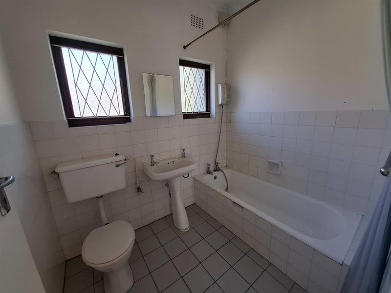 A dated bathroom featuring a pedestal basin, toilet, and bathtub with a wall-mounted electric shower unit, finished with white wall tiles and floor tiles.