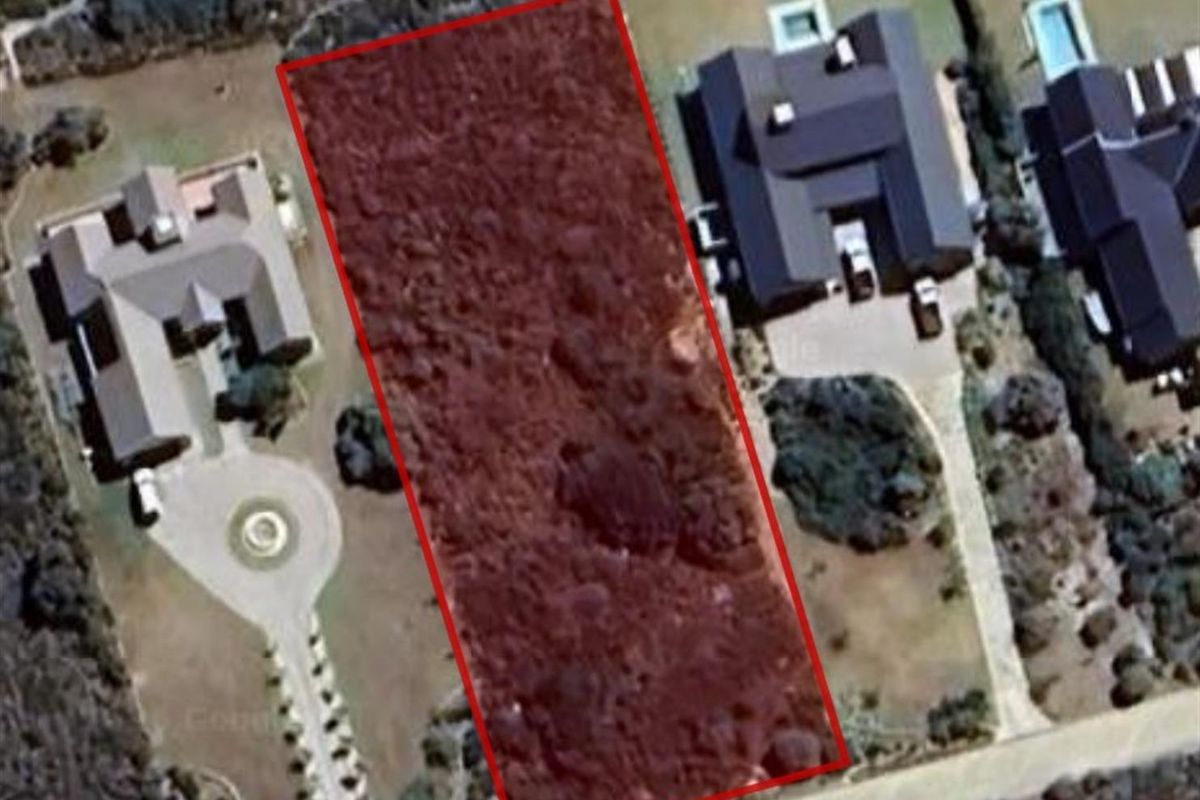 An aerial view of a vacant residential plot highlighted with a red boundary, situated between existing developed properties.
