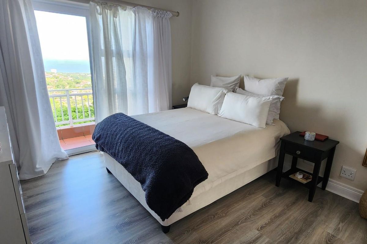 A bright bedroom featuring modern wood-look flooring, neutral walls, and a sliding glass door providing access to a private balcony with scenic views.