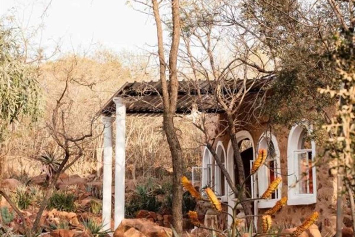 A rustic stone-walled building featuring arched windows and a covered patio area, nestled within a natural, rocky, and landscaped garden setting.