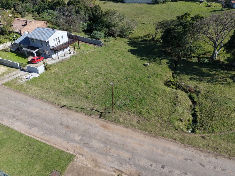 An aerial view of a vacant residential plot featuring natural grass cover, a drainage ditch, and existing boundary fencing.