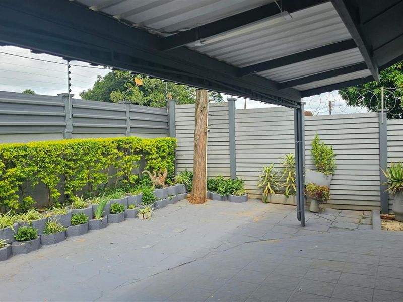 A covered outdoor patio area featuring paved flooring, a metal roof structure, and perimeter fencing with established garden beds.