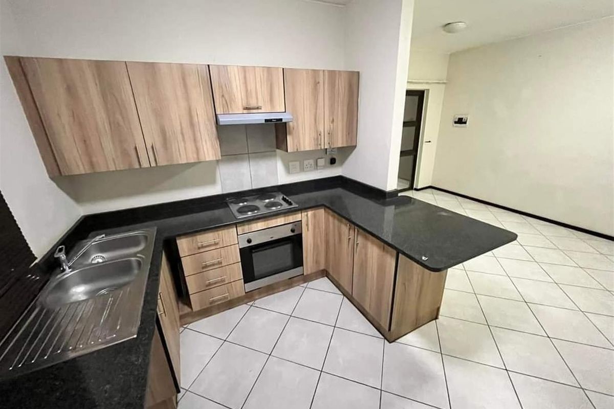 A clean, functional kitchen featuring light wood-finish cabinetry, dark countertops, built-in oven and hob, and neutral tiled flooring in an open-plan layout.
