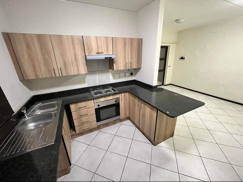 A clean, functional kitchen featuring light wood-finish cabinetry, dark countertops, built-in oven and hob, and neutral tiled flooring in an open-plan layout.