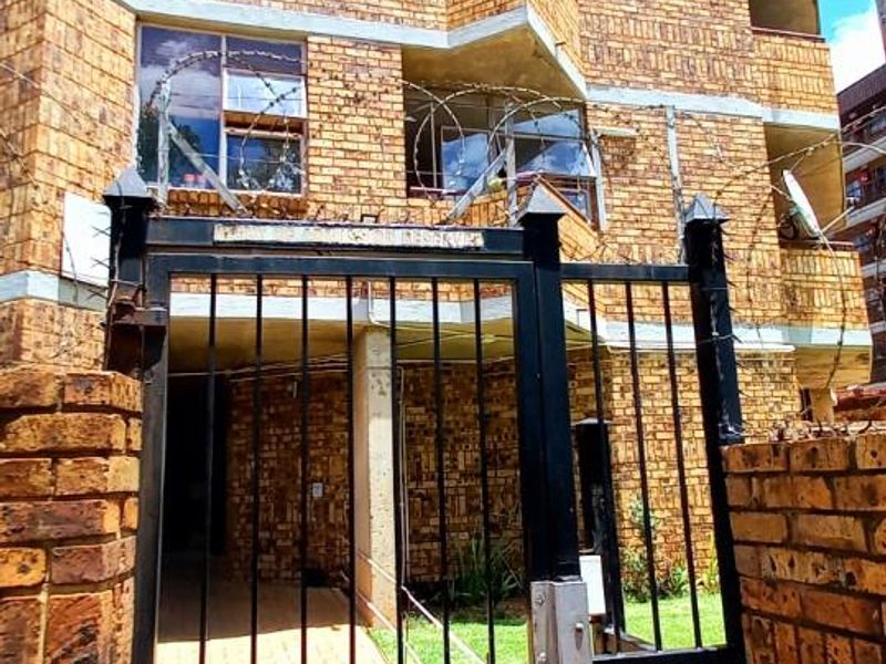 Exterior view of a multi-story brick apartment building featuring a secure metal pedestrian gate, paved walkway, and private balconies with security fencing.