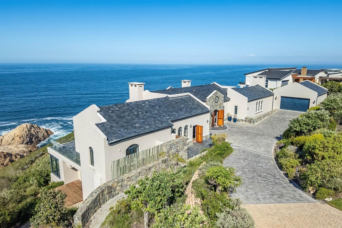 Aerial view of a luxury coastal residence featuring stone cladding, slate roofing, a paved driveway, and expansive ocean views.