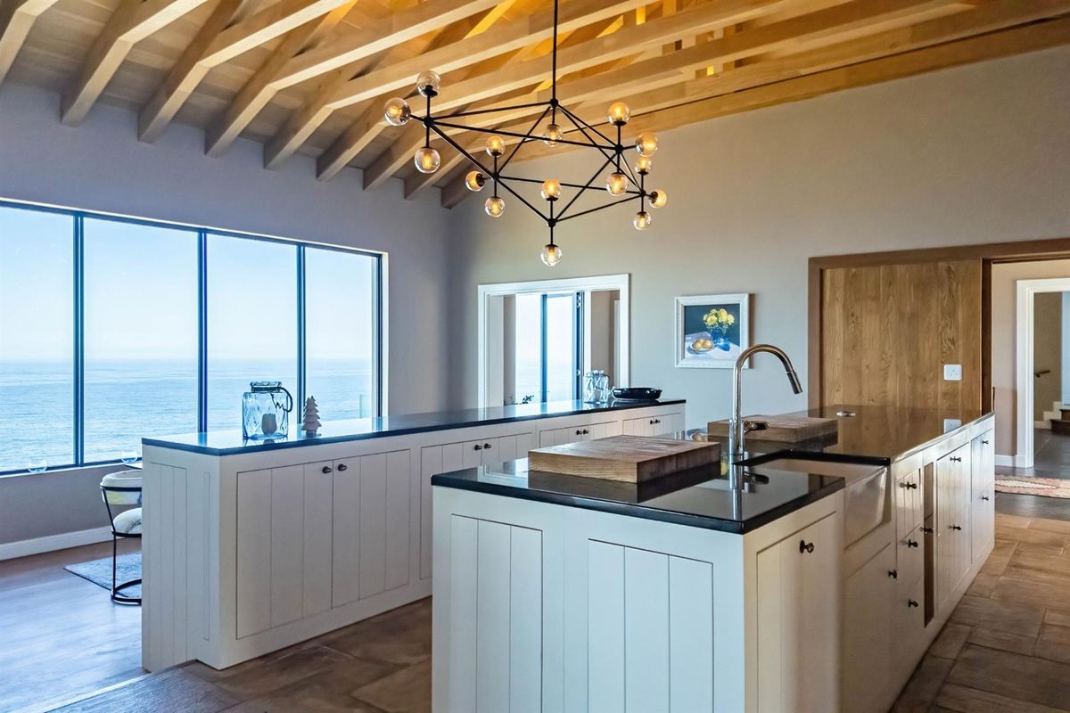 Modern kitchen featuring custom cabinetry, stone countertops, a farmhouse sink, and exposed timber ceiling beams, complemented by expansive ocean views.