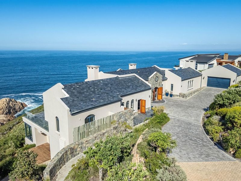Aerial view of a luxury coastal residence featuring stone cladding, slate roofing, a paved driveway, and expansive ocean views.